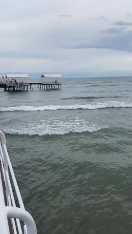 The video shows a view of a calm sea with gentle waves under a cloudy sky. Two piers with white shelters are visible in the distance, with a few people seated under them. The water appears somewhat murky green. The perspective is from a white railing, suggesting the viewer is on a boat, ferry, or another pier. The scene is tranquil, with the overcast weather creating a subdued atmosphere. The mountains are faintly visible on the horizon. The time of day appears to be late afternoon or early evening, given the soft light. The location is identified as Choktal, Kyrgyzstan, implying a freshwater lake environment rather than a saltwater sea.