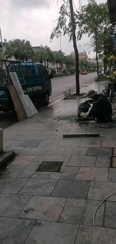 The video depicts a worker cutting metal on a sidewalk next to a road. The scene takes place on a cloudy, possibly rainy day, as the pavement appears wet. The worker is crouched down, using a grinding tool that produces sparks as it cuts through the metal. To the left of the worker is a small blue pickup truck with a ladder rack. Leaning against the truck is a sheet of material, possibly wood. In the background, a road stretches into the distance, with cars passing by. Trees and buildings line the road. The sidewalk is paved with gray tiles.