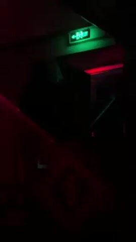The video depicts a dimly lit indoor space with red and green lighting. A green exit sign illuminates the upper right of the frame. Silhouetted figures appear to be ascending stairs, and there is a symbol resembling a section mark visible on a red surface near the bottom of the frame. The environment is dark, making details hard to discern, but there seems to be a doorway near the top of the stairs. There are sounds of music in the background.