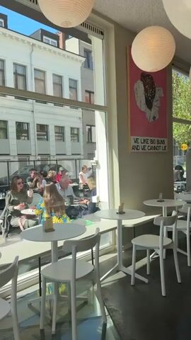 The video shows the interior of a bright cafe or restaurant with large windows overlooking a city street in Antwerp, Belgium. Inside, there are several white tables with matching chairs, some of which are empty while others are occupied by patrons. White spherical paper lanterns hang from the ceiling, contributing to the bright and airy atmosphere.

Through the large windows, an outdoor dining area is visible, where several people are seated at tables, socializing and enjoying the sunny weather. Beyond the outdoor seating, a street scene unfolds with classic European-style buildings, parked cars, and more pedestrians. The time of day appears to be daytime, with strong sunlight casting shadows, suggesting clear weather.

On an interior wall, a large framed poster is prominently displayed. The poster features an illustration of what appears to be a pastry or bun, with text below that reads, "WE LIKE BIG BUNS AND WE CANNOT LIE," adding a playful and quirky touch to the decor. The overall scene depicts a lively and inviting cafe environment where people are relaxing and enjoying their day.