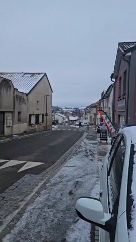 The video shows a street in a snow-covered town. The street slopes downward and is lined with houses on both sides. Some houses have snow on their roofs. There is a person walking down the street towards the camera. A 30 speed limit sign is visible. A car is parked on the side of the road in the foreground, and a pizza sign is next to it. In the distance, there is a snowy landscape with a few buildings and some power lines. The sky is overcast.