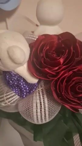 A close-up shot focuses on a floral arrangement. The primary elements are deep red satin roses with layered petals. These roses are surrounded by white mesh with vertical stripes and purple accents, possibly ribbon or fabric with polka dots. A small, white, abstract figurine, resembling a rabbit or a snowman, is visible at the top of the arrangement. In the background, a light-colored wall and a hint of a blue and beige object are discernible. The lighting is dim, creating a warm glow.