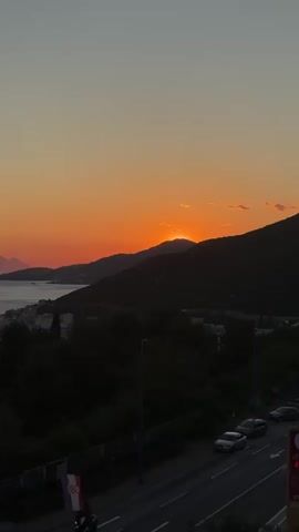 The video shows an orange sunset over a landscape of mountains, trees, and water.  The sun is setting behind the mountains, casting an orange glow in the sky.  A few clouds are present in the sky. A body of water reflects the orange glow. In the foreground, there is a road with a few cars driving on it. Trees line the roadside. There is a Croatian flag in the lower left corner. Streetlights are also visible. A bridge connects two landmasses over the water. Buildings and houses are nestled among the trees near the water.