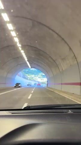 The video begins from the perspective of someone in a car, looking out the windshield. The car is traveling through a tunnel with lights lining the ceiling. The tunnel walls appear to be made of concrete. As the car exits the tunnel, the view opens up to a highway with mountains and greenery in the background. The sky is blue with some clouds. Other cars are visible on the road. The car continues on the highway, with the landscape of mountains and trees continuing to be visible on either side of the road.