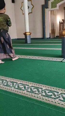 A young boy wearing a black cap, a green long-sleeved shirt, and black patterned pants walks on a green carpet inside a building. The carpet has white and green patterned borders. The boy walks from left to right in the frame, then turns to face left and stops briefly before continuing to walk away from the camera. There are white pillars with gold accents and patterned walls visible in the background.