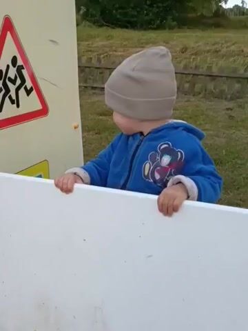 The video shows a young child wearing a grey beanie and a blue jacket with a teddy bear design. The child is standing behind a white barrier, looking towards a large, light-colored panel on the left. On the panel, there is a triangular warning sign with a red border and a black silhouette of two figures running. Below that, a yellow and blue triangular sign is partially visible. The child's hands are resting on the white barrier, and they appear to be looking intently at the signs. The background shows a grassy area with some trees and a wooden fence. The lighting suggests it is daytime, likely afternoon, with no strong indication of weather conditions other than daylight. The overall scene appears to be an outdoor educational or play area, possibly in a park or schoolyard. The child's gaze and posture suggest curiosity or engagement with the informational signs. No other people are visible.