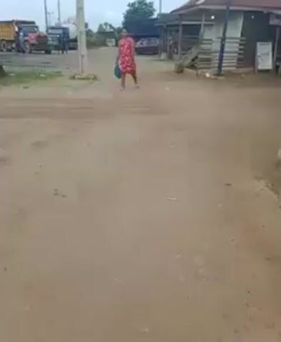 A woman in a red and white floral dress and sandals walks towards the camera on a dirt road. She carries a blue bag. Behind her are several large trucks and a small, wooden structure with a blue roof. Overhead are power lines. The sky is overcast.