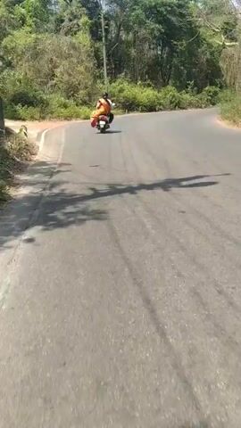 The video depicts two people riding a scooter on a curving road. The person in the back wears a bright orange outfit. The camera follows behind the scooter as it navigates the road, which is lined with trees on both sides. Another scooter appears in the distance heading in the same direction. The environment is sunny, and shadows are visible on the road.