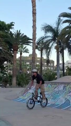 A person on a BMX bike rides on a concrete ramp in a park. The ramp has graffiti art on it. The person is wearing a black t-shirt, khaki cargo pants, white shoes, and a white baseball cap. They maneuver the bike on the ramp with a focused expression. A few other people are visible in the background, sitting and watching. The environment includes trees, grass, and an urban setting. The weather appears to be mild with overcast skies.