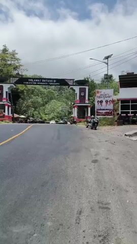 The video shows a view of a road leading towards an archway that says "SELAMAT DATANG DI KABUPATEN MINAHASA TENGGARA". The archway has red and white decorations and is flanked by pillars. There are trees and greenery on either side of the road. A few motorcycles and a car are visible near the archway. There is a poster on the right side of the archway with images of people. The road is paved and has a yellow line marking. The sky is cloudy. The video appears to be filmed from a moving vehicle.