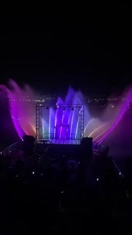 This is a nighttime video of a water fountain show. The fountains are illuminated with colored lights, primarily purple and pink, with some yellow and orange on the sides. The water jets are arranged in a semi-circular pattern behind a raised stage. The stage has a structure on it with lights and a small platform. In the foreground, there are silhouettes of people watching the show. The background shows a dark sky with some distant city lights. The video is shaky, suggesting it was recorded handheld.