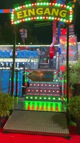 The video shows the entrance to an amusement park ride. A large sign with the word "EINGANG" (German for "entrance") in illuminated letters is visible at the top. Below the sign, a staircase is lit with colorful flashing lights. An arrow with "EINGANG" also points towards the stairs. The ride itself appears to be a thrill ride with multiple rows of blue seats. The background features graphics related to motocross, with images of motorcycles and riders performing stunts, and the text "NIGHT OF FREESTYLE." The scene is dimly lit, suggesting it is evening or night, and there are potted plants on either side of the entrance ramp. There is at least one person visible on the ride, seated in one of the chairs. The ride is stationary at the moment.