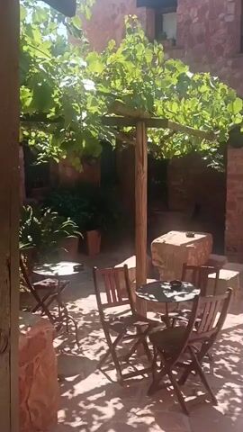 The video shows a shaded outdoor patio area with a table and chairs set up under a grape arbor. The patio is paved with light-colored stones, and the walls of the surrounding building are made of red brick. In the background, there is a doorway leading into the building, and a window with a red shutter. The sun casts dappled shadows on the patio, indicating it is daytime. There is a potted plant to the left of the doorway, and some greenery to the right. A large, rough-hewn stone block is positioned near the table. The scene appears to be serene and inviting.