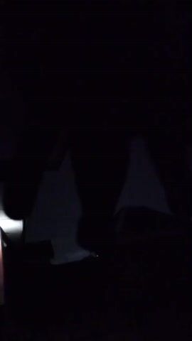 The image displays a very dark, outdoor scene, likely at night. Two prominent dark, triangular shapes resembling tents or temporary shelters are silhouetted against a slightly lighter background in the mid-ground. On the left side of the frame, a distinct, bright light source, possibly a flashlight or lantern, casts a localized glow, partially illuminating a small area in the foreground. Below the main tent-like structures, other indistinct dark forms are visible, blending into the dark surroundings. No people are visible. The extreme darkness and reliance on an artificial light source confirm the time of day as night. The setting suggests a camping scenario or a temporary outdoor arrangement in Ilogbo Eremi, Nigeria. There is no visible text.