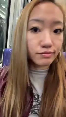 A woman with long, light brown hair sits on public transit. She is wearing a maroon jacket over a white shirt with a design. She has silver hoop earrings in and purple earbuds. She looks at the camera. Other passengers are visible in the background.