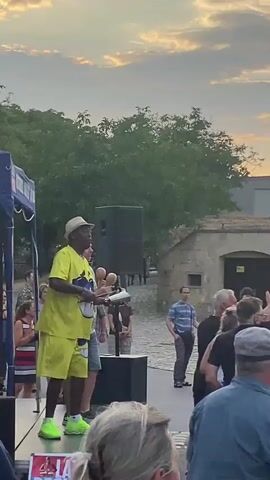 A man in a yellow t-shirt, yellow shorts, and a white hat is standing on a stage holding maracas. He is in front of a crowd of people. To his left, there is a blue canopy and a speaker. To his right, there is another speaker and a stone building. The sky is cloudy and the sun is setting. A group of people are dancing in front of the stage. The man is shaking the maracas.