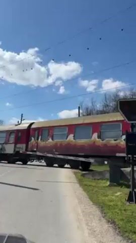 The video shows a train crossing a road on a sunny day. The train consists of older, red passenger cars with graffiti. The railroad crossing has a red light and a lowered gate. A car is waiting on the other side of the tracks. The sky is blue with scattered white clouds. There are overhead electrical wires. Trees line the area beyond the tracks. The camera seems to be filming from inside a car waiting at the crossing.