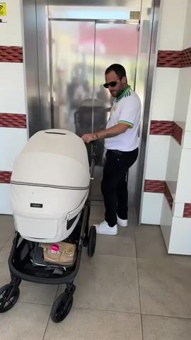 A man with a beard, dark glasses, a white t-shirt with a gold and green collar, black pants, and white sneakers stands next to a baby carriage and faces an elevator. The baby carriage is white with a black base. The man looks at his phone and then at the baby carriage. The elevator doors have a reflective surface that shows a woman holding a baby in the background. The man has tattoos on his arms. The elevator is in a hallway of some sort that has a white, brown, and silver wall.