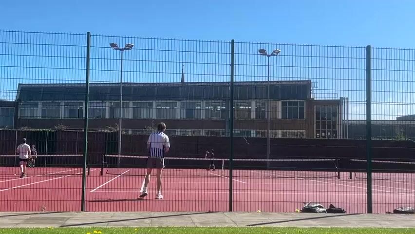 Two men are playing tennis on a red hard court on a clear, sunny day in Dundee, United Kingdom. The camera is positioned behind a green wire mesh fence, looking towards the court and a large building in the background. The main subject, a man in a white and blue striped shirt and light shorts, is seen from behind, actively participating in a rally. He is captured jumping to hit the ball with his racket around the 0:01 mark and then returning to a ready stance. Another player, dressed in a white shirt and dark shorts, is visible on the left side of the court. A third person in dark attire is also present on the far side of the court. The court has white boundary lines and a net. In the background, under a bright blue sky, stands a large, multi-story building with numerous windows, possibly a school or university, and a small spire on its roof. Two tall light poles with multiple lamps illuminate the court. Two sports bags and some tennis balls are visible on the ground beside the fence to the right.
