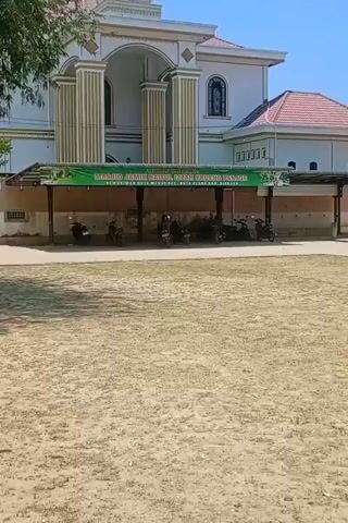 The video shows the exterior of two buildings, a parking area, and a man. The camera pans from left to right, starting with a two-story white building with gold-painted columns and arched windows on its upper floor. A green banner with white text is strung across a covered parking area filled with parked motorbikes. In the foreground, there is a sandy area with sparse vegetation.

As the camera continues to pan, a man wearing a yellow shirt and dark pants walks up a set of steps towards a gray, unpainted building. This building also has arched windows. The sky is clear and blue, and some tree branches with yellow flowers hang down from the top of the frame.