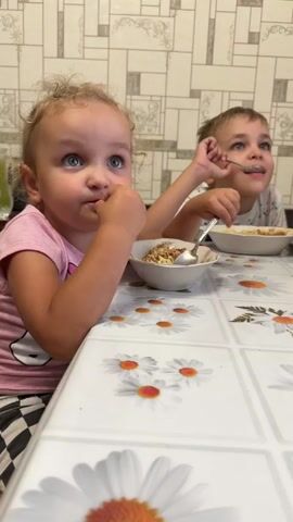 Two young children, a girl and a boy, are sitting at a table eating. The girl, in the foreground, is wearing a pink t-shirt with a unicorn design and black and white checkered pants. She has blonde hair in pigtails and is looking up with her mouth open, her fingers near her mouth. The boy, in the background, is wearing a white t-shirt and is eating with a spoon, looking to his right. Both children have bowls of food in front of them, and the table is covered with a white tablecloth decorated with daisies. The background appears to be a kitchen with tiled walls. The lighting suggests it could be daytime. There is a calendar visible on the wall in the background with dates up to 2026. The scene depicts a casual mealtime.