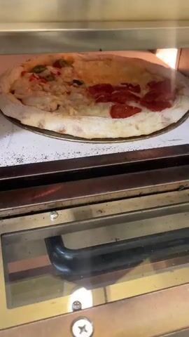 A split pizza, half pepperoni and half with chicken and jalapeños, is baking in an oven. The pizza is on a metal baking sheet, which rests on a rack inside the oven. The oven interior is visible, with a heating element glowing at the top right. The front of the oven has a metal handle and a glass window, through which the pizza is being viewed. The pizza appears to be nearly cooked, with the cheese beginning to melt and bubble. The crust is rising and browning. The overall scene suggests the pizza is in the final stages of preparation.