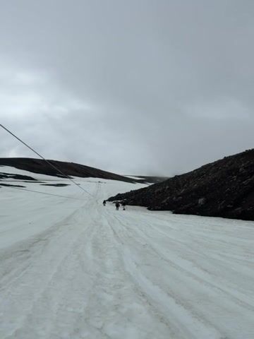 The video shows a group of people walking on a snow-covered mountain. The terrain is rocky and dark on the right side of the frame, with a cable running diagonally across the left side. The sky is overcast and gray. Several individuals are visible in the distance, walking up the snowy slope. Closer to the camera, four people are walking, one in a blue jacket and red backpack, another in a pink jacket, and one person in a yellow jacket. One person appears to be using trekking poles. The overall impression is of a challenging hike in a cold, possibly remote environment.