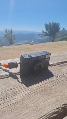 The video shows a black compact camera placed on a weathered wooden table. The camera has a strap attached, with an orange bead and a black connector. In the background, there is a dry, grassy landscape with scattered trees and rolling hills under a clear blue sky. Sunlight beams down from the upper left corner, creating a lens flare. Towards the end of the video, a plastic water bottle and part of a black backpack are visible on the table.