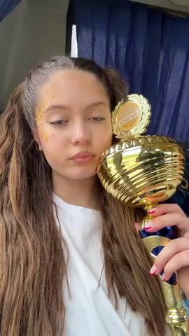 A young woman with glitter on her forehead holds up a gold trophy that reads "Artist Hub 2024." She is on what appears to be a bus with blue patterned seats. The woman is wearing a white t-shirt, has her brown hair in pigtails, and is making different facial expressions at the camera, including winking and puckering her lips. She is holding the trophy with one hand and occasionally moves it around to show more of the trophy. She is wearing several bracelets. The bus has blue curtains behind her.