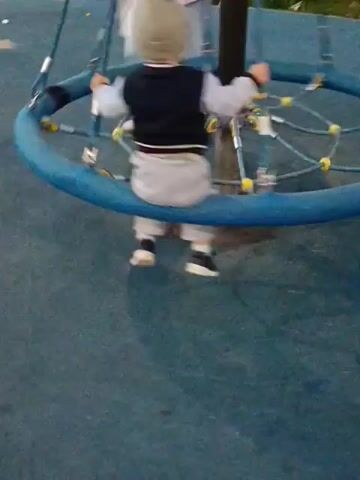 A woman and a toddler are on a playground merry-go-round. The woman, wearing a pink jacket and light-colored pants, stands on the outside of the merry-go-round and appears to be pushing it. The toddler, wearing a grey hat, a dark vest over a white shirt, and grey pants, is sitting in the middle of the merry-go-round and holding onto the central pole. The merry-go-round is blue and has ropes attached to it. The ground around the merry-go-round is blue. In the background, there are buildings, cars, and some greenery. The scene appears to be outdoors in the daytime.