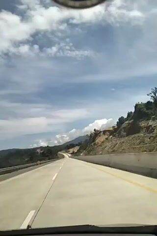 This video captures a first-person perspective from inside a vehicle traveling on a highway in Lamtamot, Indonesia. The scene is dominated by a multi-lane highway with a clear blue sky above, dotted with scattered white clouds. The highway is paved with concrete and marked with white and yellow lines. To the right of the highway, there is a steep, rocky hillside covered in sparse vegetation and trees. Beyond the hill, more distant mountains are visible, some with white cloud formations around their peaks. To the left of the highway, the landscape is greener with more dense trees. The time of day appears to be daytime, likely late morning or afternoon, given the bright sunlight casting shadows. The weather seems fair with a mix of sun and clouds. There is no visible text in the video. The overall mood is one of peaceful travel through a scenic, mountainous landscape.