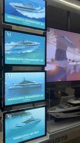 The video shows a display featuring multiple screens and yacht models. On the left, a vertical stack of four illuminated screens displays images of different Mangusta yachts, each with the model name printed below: Mangusta Gransport 34, Mangusta Gransport 38, Mangusta Gransport 50, and Mangusta 165 Rev. To the right of these screens, a large horizontal screen displays a video of a luxurious yacht deck with an infinity pool and a person in the water. Below the large screen, a detailed model of a yacht is showcased within a glass display case. The overall environment appears to be a showroom or exhibition space, with branding for "MANGUSTA" visible on the display. The lighting is soft and highlights the products.