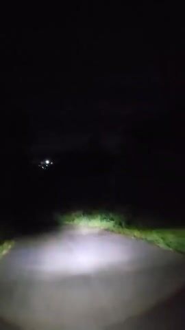 The video shows a dark scene illuminated by a single light. A narrow paved road or path stretches forward, flanked by green grass or vegetation on either side. Beyond the illuminated area, the background is almost completely dark, with indistinct shapes of trees or other foliage barely discernible. The perspective suggests a point of view from a low position, possibly a vehicle or a person holding a light source close to the ground. The overall impression is one of nighttime or a very dimly lit area, with the single light creating a tunnel-like view.