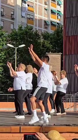 A group of people, mostly elderly women wearing white t-shirts and black pants, are performing a synchronized dance routine on a wooden stage outdoors. A younger man in a white t-shirt and black shorts with white stripes appears to be leading the dance. The background shows a brick building with several floors and balconies, trees, and a street light. Some audience members are visible in the foreground, with one holding up a smartphone. The overall atmosphere appears to be a lively outdoor event.