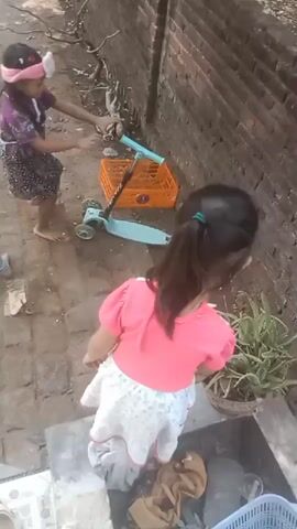 Two young girls are playing outdoors on a paved area next to a brick wall. The girl on the left, wearing a pink and purple outfit and a pink headband, is standing on a blue and green scooter. She is reaching forward with her right hand. The girl on the right, wearing a pink shirt and a floral skirt, is sitting on a concrete ledge facing away from the viewer. In front of the scooter is an orange plastic crate. To the right of the girls, there is a potted plant. The ground is made of uneven pavers. There is also some laundry on the ledge where the second girl is sitting.