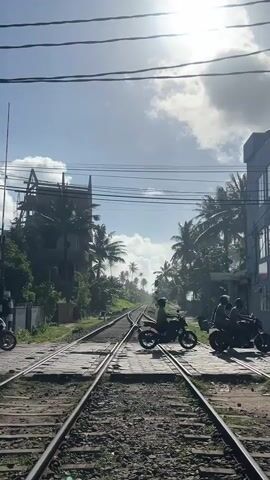 This vertical video depicts a sunny day in Kodagoda, Sri Lanka, with a view of railway tracks stretching into the distance. Several people on motorcycles are waiting at a railway crossing. The sun is bright, casting strong highlights and shadows, suggesting it is midday. Palm trees line the sides of the tracks, and in the background, there are buildings, including one under construction. The overall atmosphere appears casual, with people waiting their turn. The railway crossing is made of wooden planks. The sky is blue with some scattered clouds. There is no visible text in the video.