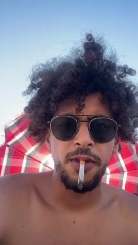 The video shows a close-up of a person's face. They have curly dark hair, sunglasses, and a beard. They are smoking a cigarette. In the background, a red and white checkered umbrella is visible against a clear blue sky. The person in the video gestures with their middle finger towards the camera.