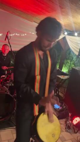 A man in a black outfit with colorful striped suspenders plays a yellow drum. He is performing at what appears to be an outdoor event with white draped fabric overhead and red lighting. In the background, another man is playing a drum set, and various electronic equipment, including what looks like a keyboard and speakers, are visible. The overall atmosphere suggests a live music performance.