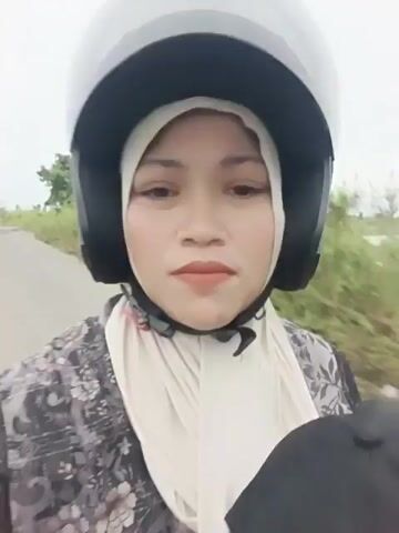 A woman wearing a hijab and a black helmet is riding on a motorcycle. She is looking directly at the camera and smiling slightly. The background is a blur of greenery, suggesting the motorcycle is in motion. There are other people on the motorcycle behind her, their faces obscured. The sky is overcast.
