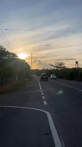 The video shows a road with cars driving during sunset. The sun is low in the sky, casting a warm glow. There are trees and vegetation on either side of the road. The road has a white line dividing the lanes.