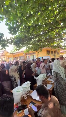 The video shows a crowded outdoor scene with many people standing in line. The environment appears to be sunny with large green trees overhead providing shade. In the foreground, there is a wooden table with several papers and what appear to be bags of rice or flour. Some people are interacting with the table, possibly for registration or distribution. The majority of the people are dressed in modest clothing, with many women wearing headscarves (hijabs). The mood seems orderly, with people patiently waiting their turn. There are some buildings in the background. The camera angle is from a slightly elevated position, looking down at the crowd and the table.
