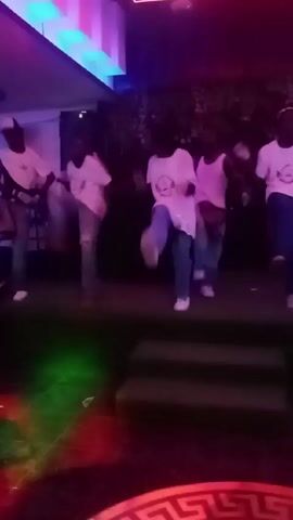 Here is a concise description of the video:

The video shows a group of five or six young people dancing on a stage under colored club lights. They are all dressed casually in light-colored tops and jeans, performing coordinated dance moves. The stage is small with steps leading up to it. The environment is a nightclub or similar venue, as suggested by the lighting and the presence of a table with a patterned surface in the foreground. The overall tone appears energetic and lively.