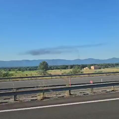 The video shows a view from a vehicle moving on a highway under a clear blue sky. A landscape with fields, scattered trees, and distant mountains is visible on the other side of the road. A thin, dark cloud of smoke hangs in the distance. A black van and a silver SUV pass by on the highway. The sun glints off the side mirror of the vehicle filming. The highway is lined with guardrails.