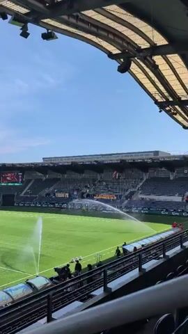 A stadium with a well-maintained green field is shown from a slightly elevated perspective. Water sprinklers are actively watering the field, creating visible arcs of water. Several people are visible on the field near the sprinklers, seemingly tending to them. In the background, tiered seating rises up, largely empty but with some spectators scattered throughout. A large video screen is mounted above the seating, displaying a team logo and score. The sky is clear and blue.