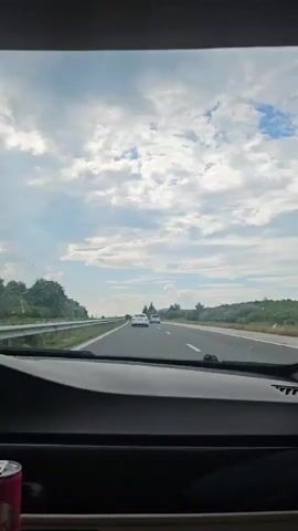 A video from a car shows a highway with cars and trees under a cloudy sky. The camera is fixed, capturing the movement of the car as it travels down the highway. The front windshield and dashboard are visible. A red and white can is visible in the bottom left corner of the frame.