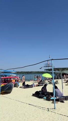 The video shows a crowded beach on a sunny day. Several people are sunbathing on the sand, some are sitting under umbrellas, and others are in the water. There are several surfboards and inflatable boats on the beach. In the background, there is a lifeguard tower with a person standing on it. The sky is clear and blue, and the water is calm.