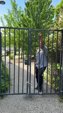A young man is seen climbing over a tall, dark gray metal gate on a sunny day in Bron, France. He wears a light-colored short-sleeved shirt, dark pants, black sneakers, sunglasses, and a black shoulder bag.

The man approaches the gate, which has vertical bars and a horizontal top bar. He first places his left foot on a small, green, box-like protrusion located on one of the gate's vertical bars. He then uses his hands to grip the top horizontal bar of the gate, lifting his body. He brings his right leg over the top of the gate, and then his left leg, carefully lowering himself to the other side. The gate appears to be secured with a lock, preventing easy passage.

In the background, there are lush green trees and foliage, indicating an outdoor, possibly park-like or residential, setting. The ground on both sides of the gate is made of light-colored gravel or concrete. The sky is bright and clear.