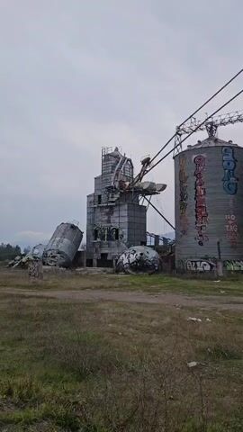 A panoramic video shows a dilapidated industrial complex with several large, cylindrical silos and a central tower. The silos, made of corrugated metal, are covered in colorful graffiti. One silo on the left is tilted at a severe angle, appearing to have collapsed or been severely damaged. Another silo lies on its side in the foreground, also covered in graffiti. A tall, multi-story concrete structure stands behind the silos, with a crane or conveyor system extending from it. The ground is covered in dry, sparse vegetation and some debris. The sky is overcast and a muted gray. The overall impression is one of decay and abandonment, with the graffiti adding a layer of artistic reclamation to the industrial ruins. The video pans from left to right, showcasing the extent of the damaged structures.