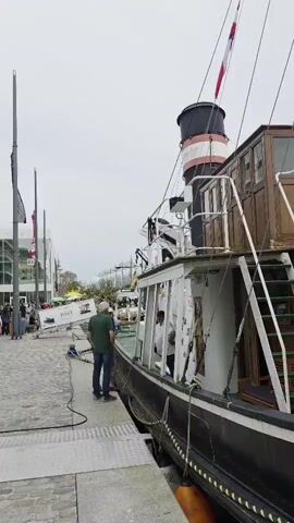A vertical video shows a waterfront scene with a tugboat docked alongside a pier. A man in a green shirt and jeans stands on the pier looking at the boat. Several other people are walking on the pier to the left of the frame. A building with a modern facade is visible behind them. The sky is overcast.
