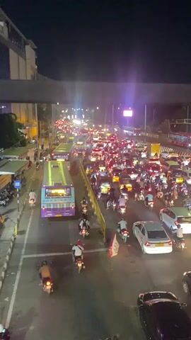 The video shows a high-angle view of a busy city street at night. Traffic is heavy, with many cars, buses, and motorcycles densely packed on the road. The street is illuminated by streetlights and vehicle headlights, and there are several tall buildings visible in the background. People can be seen walking on the sidewalks alongside the road.